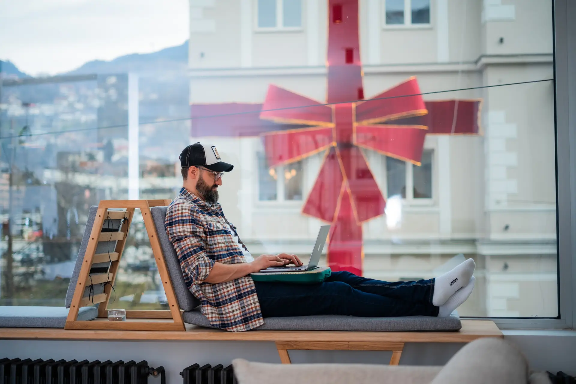 Professional sitting and working on a comfortable and aesthetic window bench in a coworking space in Sarajevo, with a laptop on his lap.