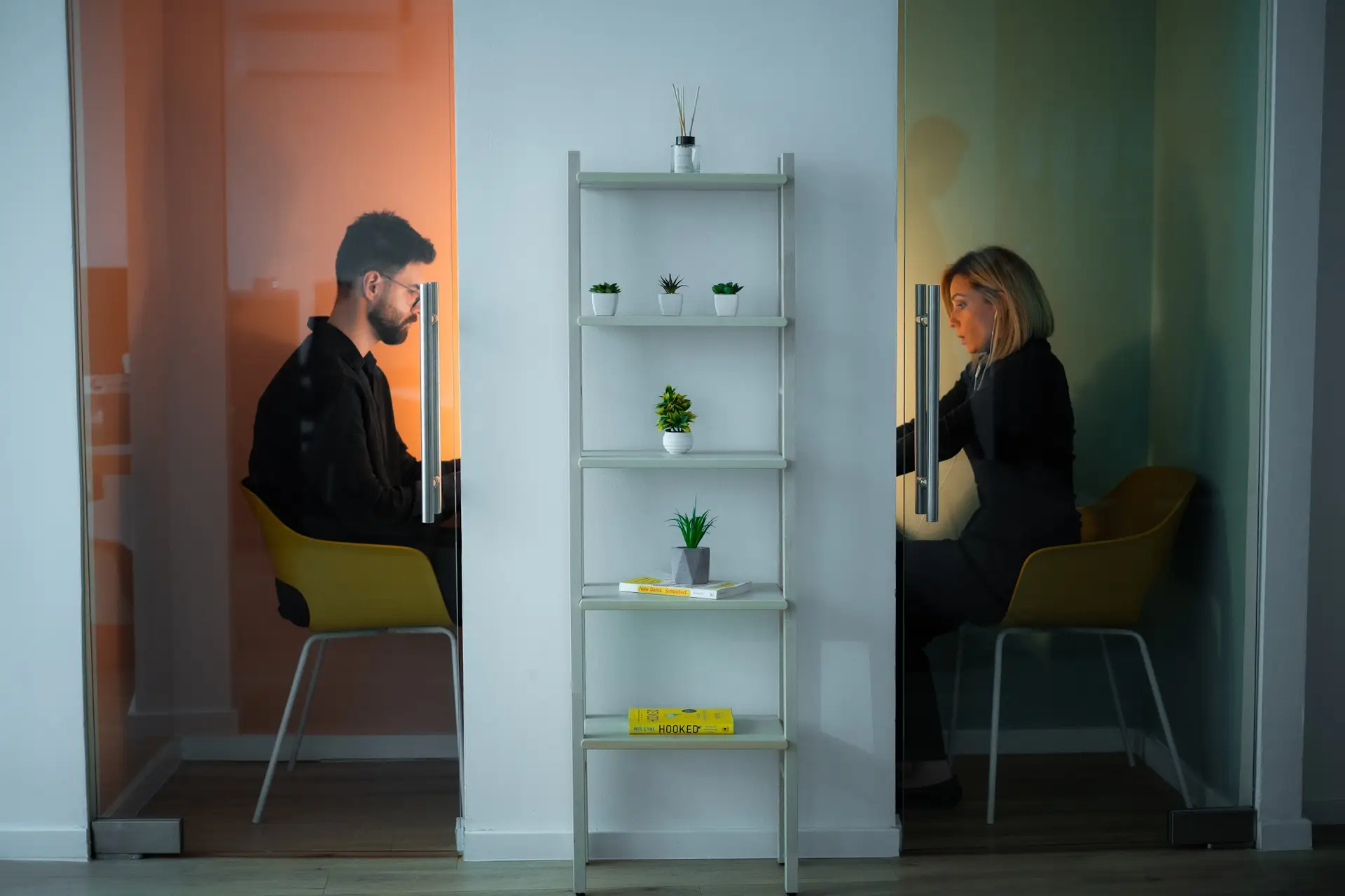 two individuals using a coworking space in Sarajevo, sitting in separate phone booths with pastel colored walls, facing each other while on private calls.