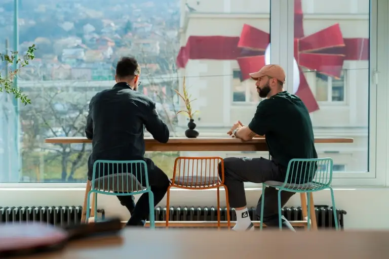 Freelancers working in coworking space in Sarajevo, seated on bar stools at a standing desk near panoramic windows with a view of Sarajevo