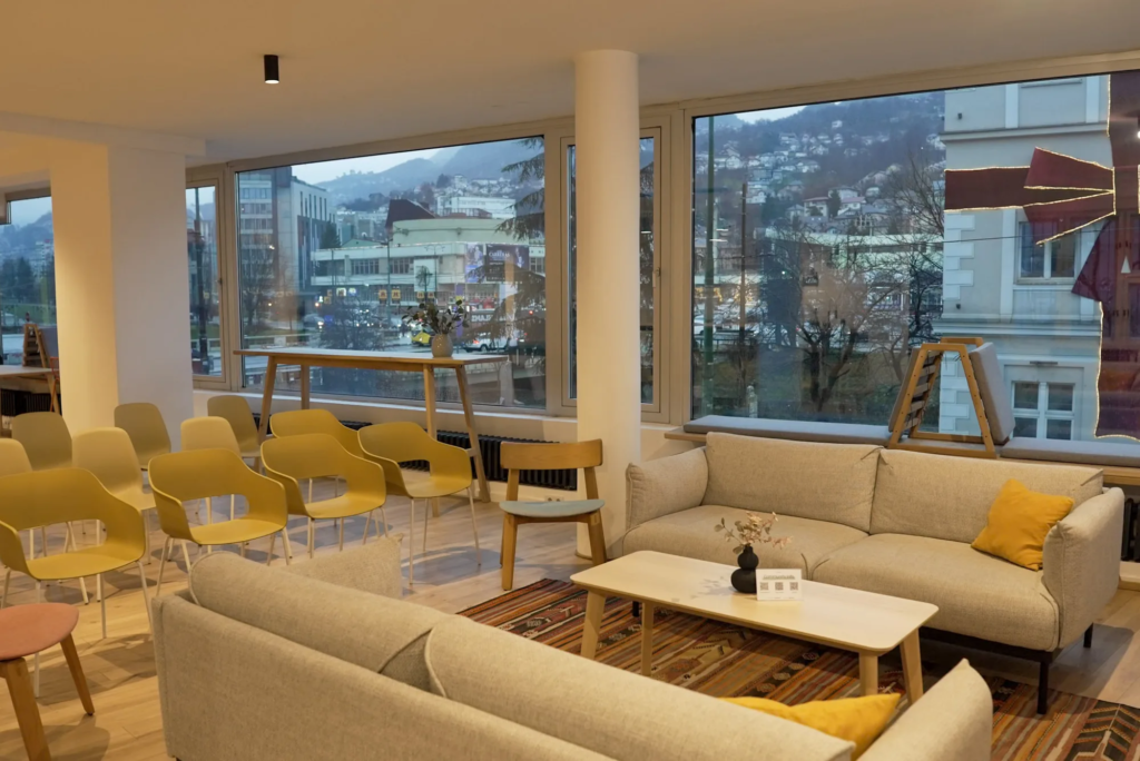 Lounge area with beige sofas, mustard chairs set up theater-style, and panoramic views of Sarajevo at dusk, Motiff coworking space